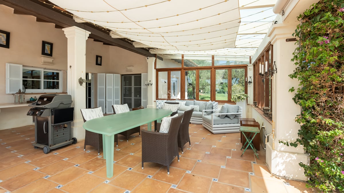 Terrasse couverte avec sol en terre cuite, table à manger, coin salon et barbecue sous un auvent en toile.