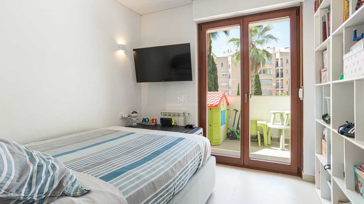 Chambre lumineuse avec lit à rayures, TV murale et baies vitrées ouvrant sur une terrasse avec jeux d'enfants.