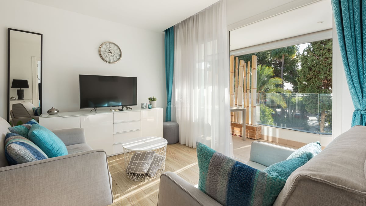 Salon moderne avec canapés gris, coussins turquoise et grandes baies vitrées ouvrant sur une terrasse arborée.