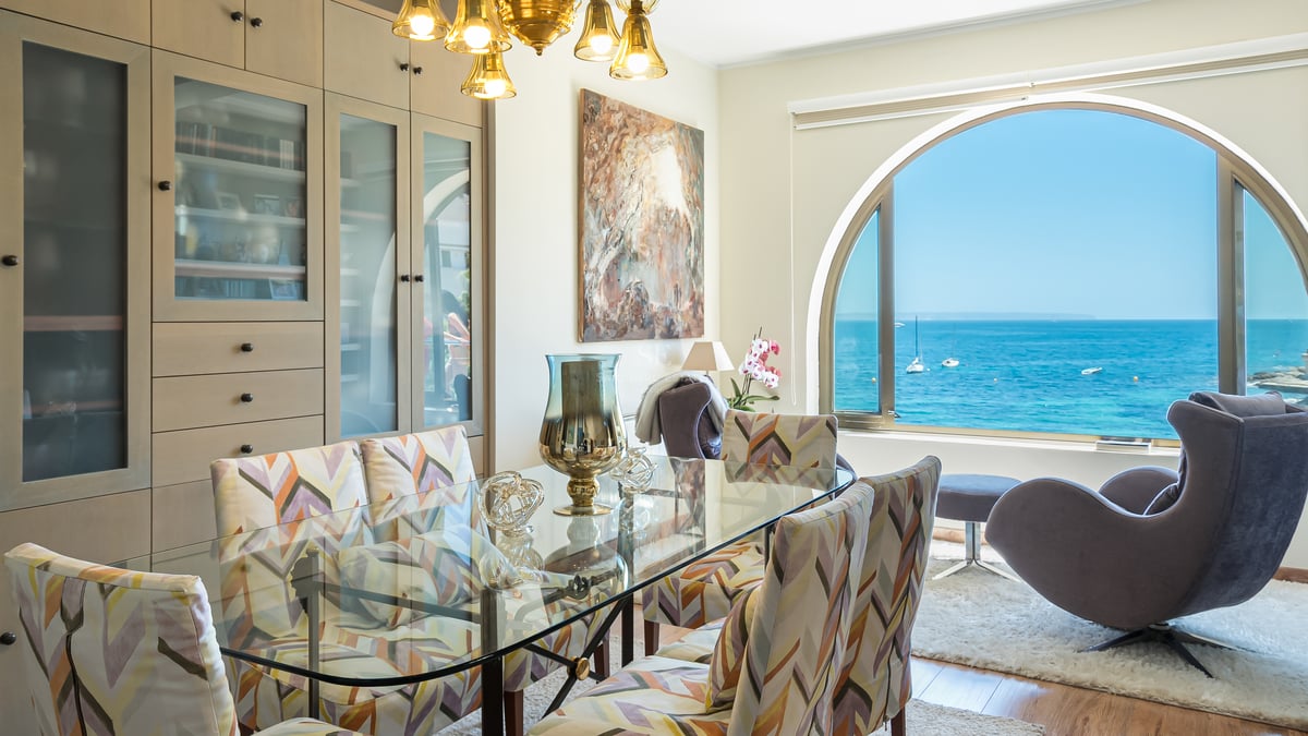 Matsal med glasbord och ett stort välvt fönster med utsikt över det blå havet och båtar.