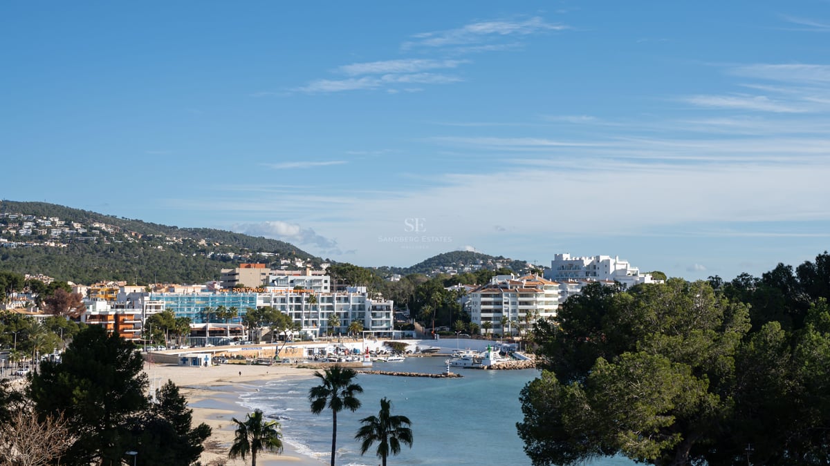 Vue dégagée sur une ville côtière avec des bâtiments blancs, une plage de sable, des palmiers et un petit port sous un ciel bleu.