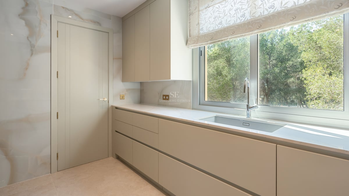 Minimalist kitchen featuring cream handleless cabinets, a large window overlooking greenery, and a stone backsplash.