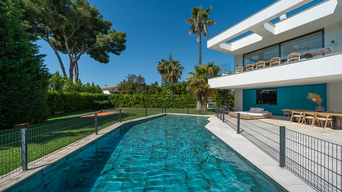 Villa blanche moderne avec piscine bleue, meubles de terrasse en bois et jardin paysager sous un ciel bleu.