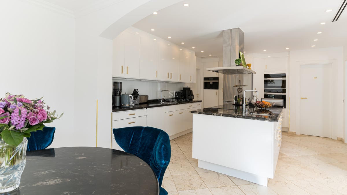 Zeitgenössische weiße Küche mit schwarzen Steinarbeitsplatten, Kochinsel und blauem Samtstuhl.