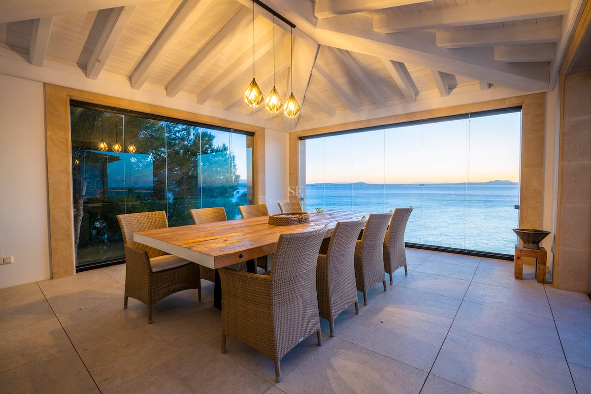 Großer Holzesstisch auf einer Terrasse mit Glasvorhängen und Blick auf das blaue Meer bei Dämmerung.