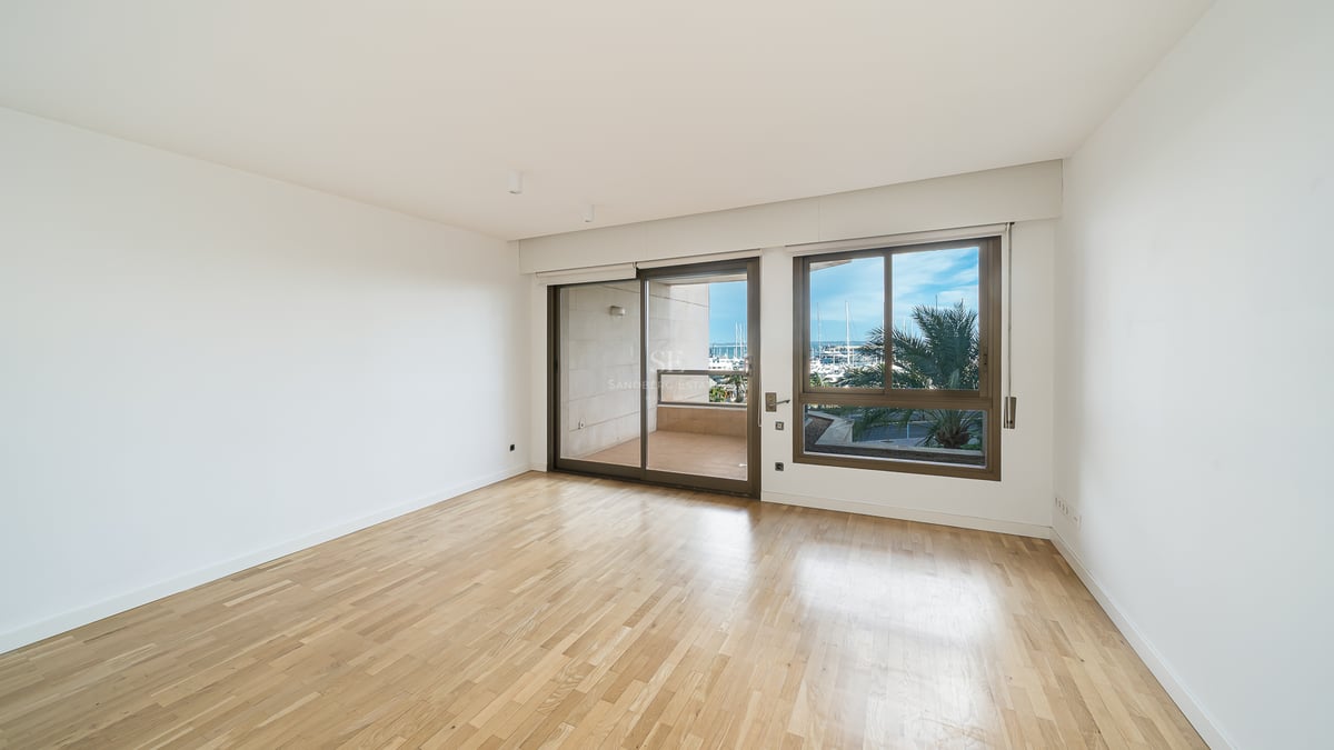 Tomt modernt rum med ljusa trägolv och stora fönster med utsikt över en balkong och en marina med palmer.