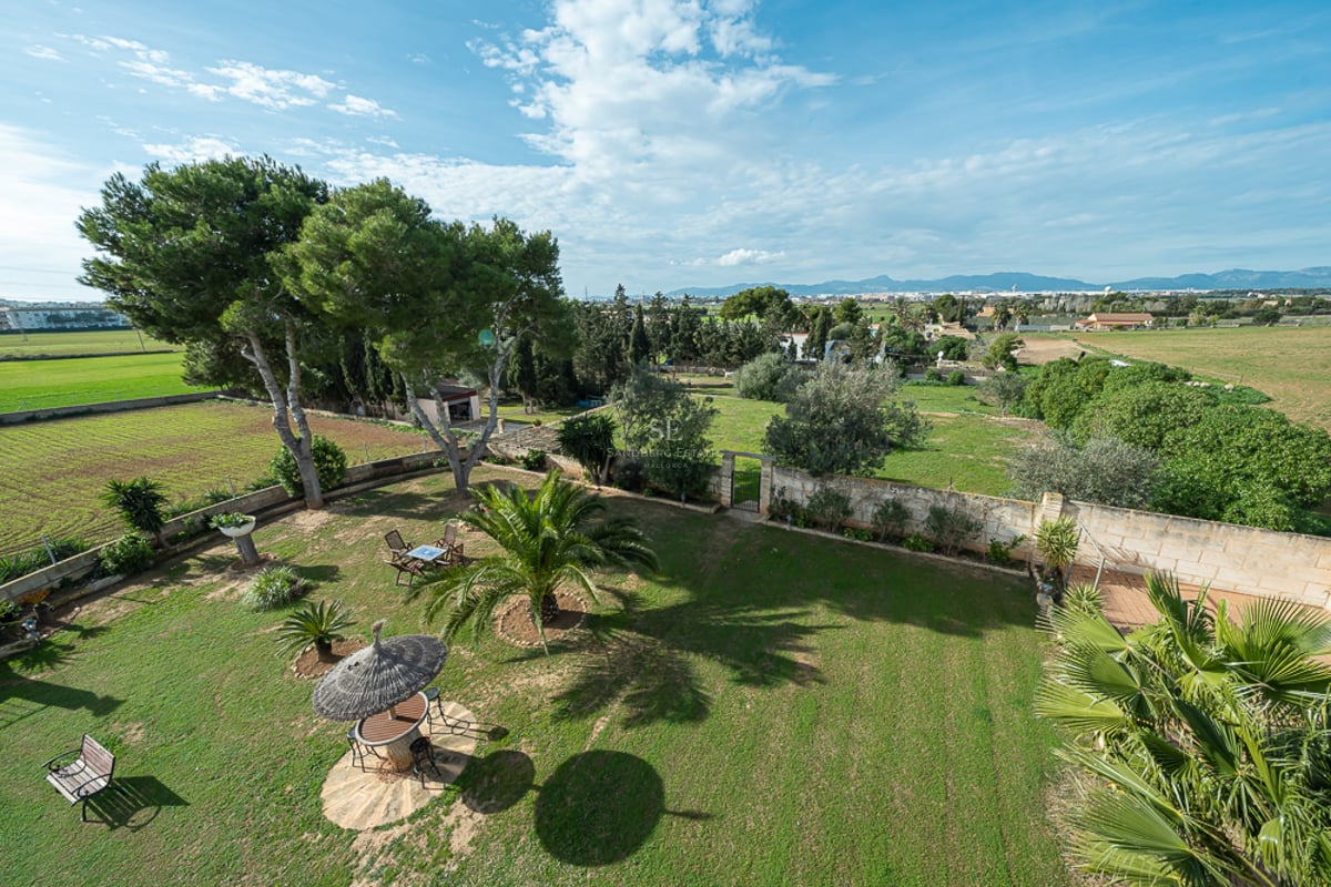 Vue surélevée d'une grande pelouse avec palmiers, murs en pierre et parasol en chaume avec les montagnes en arrière-plan.