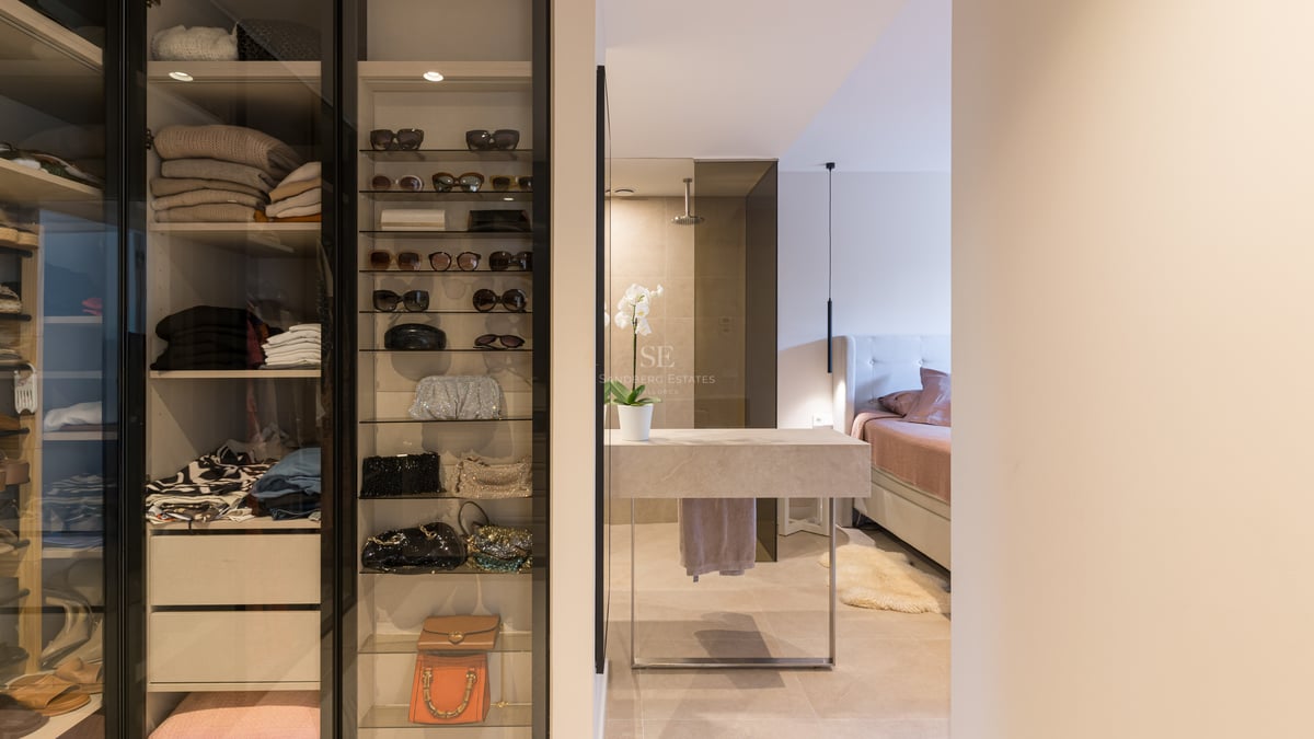 Modern walk-in closet with glass display shelves for accessories next to a stone vanity and master bedroom.