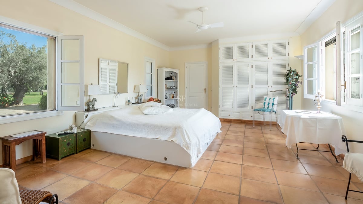 Chambre spacieuse avec sol en terre cuite, armoires à persiennes blanches et fenêtres donnant sur un jardin verdoyant.