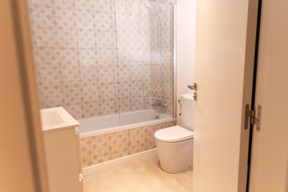 Badezimmer mit beige gemusterten Fliesen, Badewanne mit Glastrennwand, Toilette und Waschtisch.