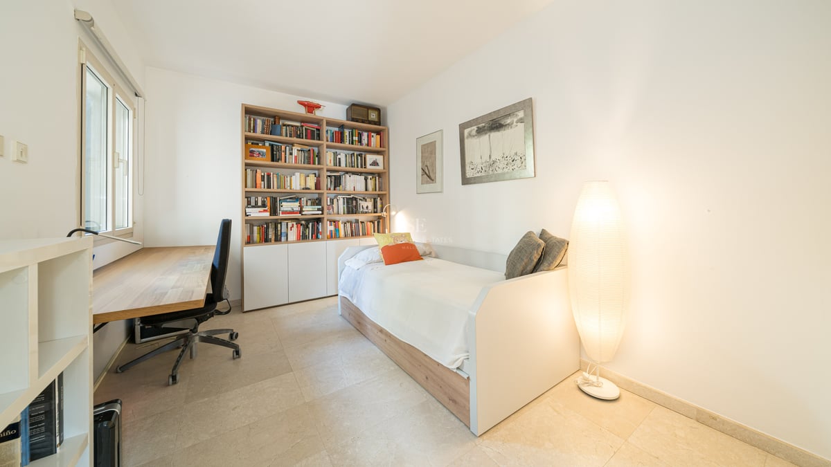 Ljust sovrum med enkelsäng, stor bokhylla i trä, skrivbord och en stämningsfull golvlampa.