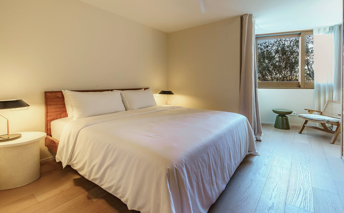 Modernes Schlafzimmer mit weißer Bettwäsche, Holzkopfteil, Designerkleuchten und einem Sessel am Fenster.