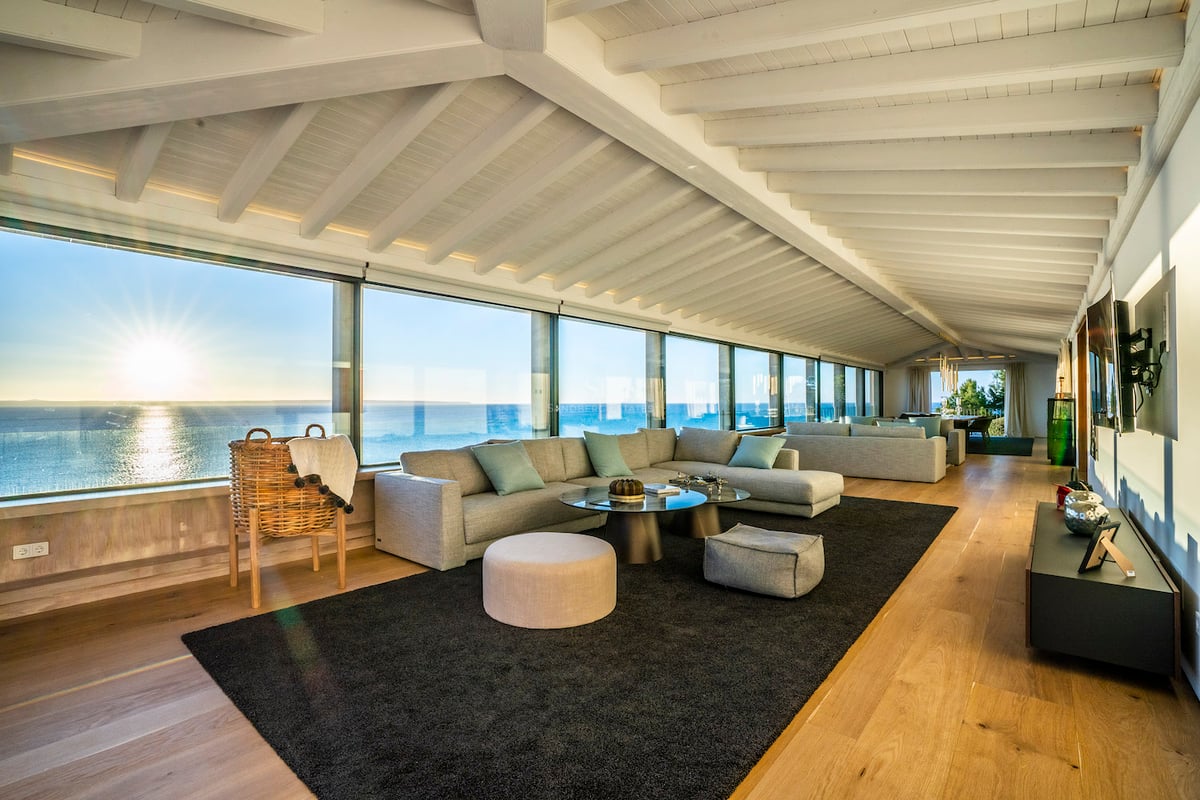 Modernes Wohnzimmer mit weißen Deckenbalken, Eichenparkett und bodentiefen Fenstern mit Meerblick bei Sonnenuntergang.