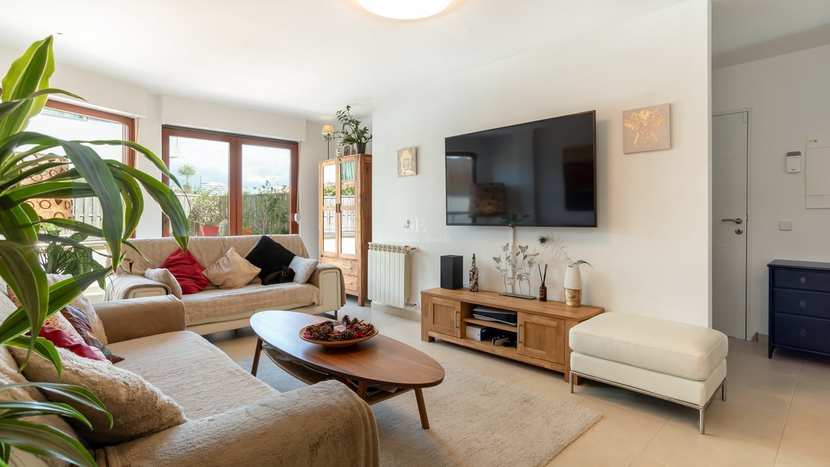 Salon lumineux avec canapés beiges, meubles en bois, grand téléviseur mural et accès à une terrasse extérieure.