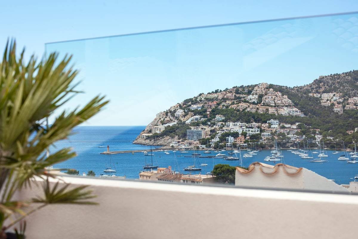 Vue sur un port méditerranéen avec bateaux et villas à flanc de colline à travers un garde-corps en verre.