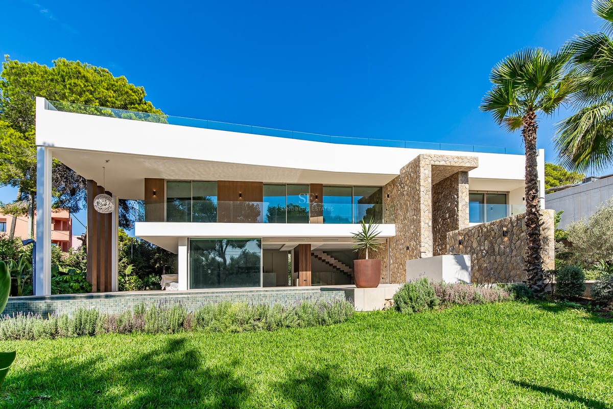 Villa moderne blanche de deux étages avec murs en pierre, balcons en verre et piscine donnant sur une pelouse verte.