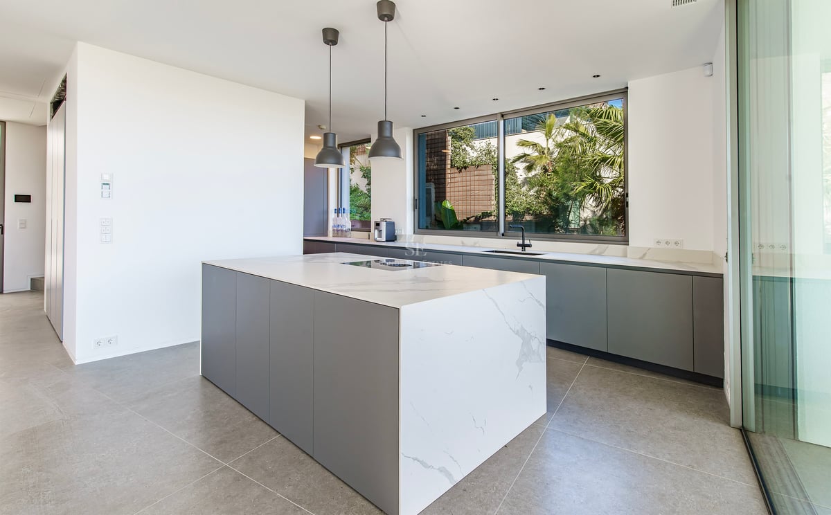 Cuisine moderne avec un grand îlot en marbre blanc, des meubles gris et une fenêtre donnant sur un jardin tropical.