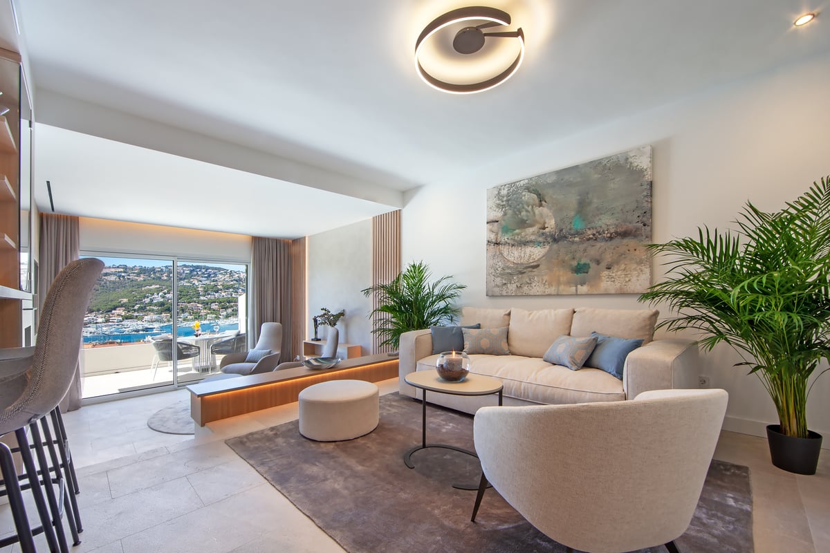 Salon moderne et lumineux avec mobilier crème, grandes baies vitrées sur la mer et plantes vertes.