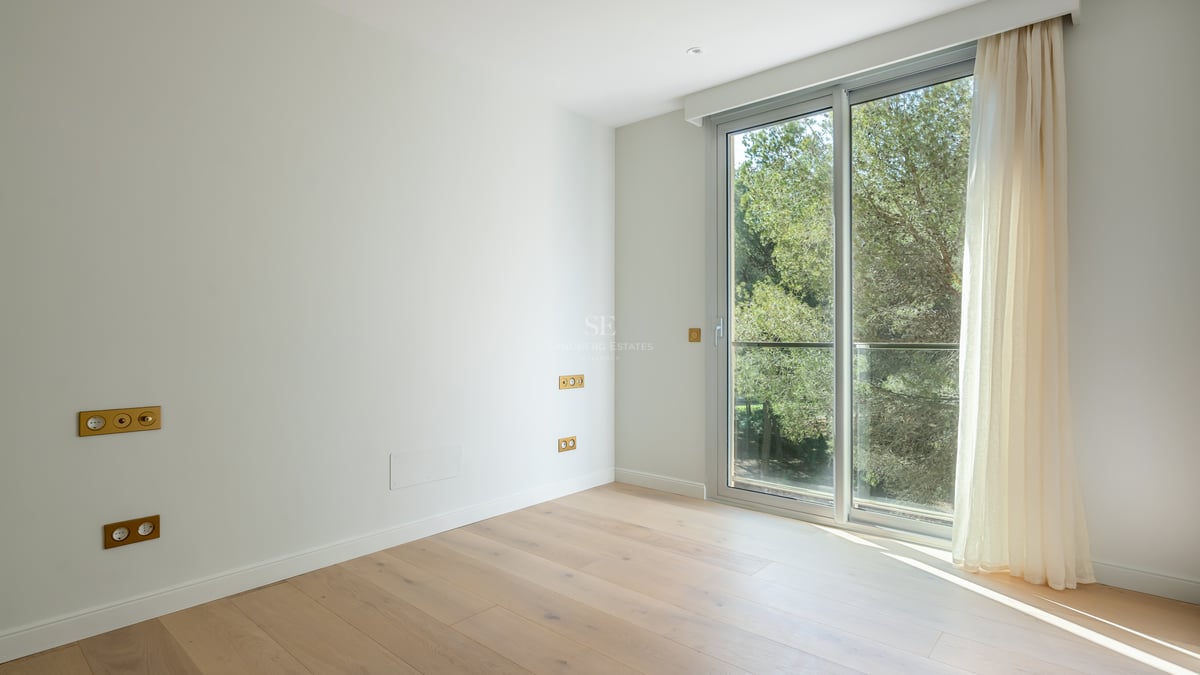 Empty modern bedroom featuring light oak floors, white walls, and large sliding glass doors overlooking green trees.