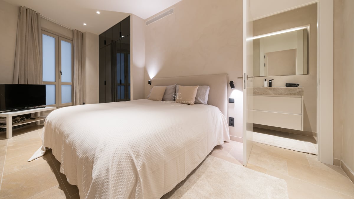 Chambre principale moderne avec textiles crème, sol en pierre, placards sombres et salle de bain attenante.