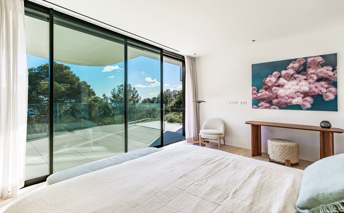 Chambre lumineuse avec lit blanc, baies vitrées du sol au plafond et grande terrasse avec vue sur les arbres.