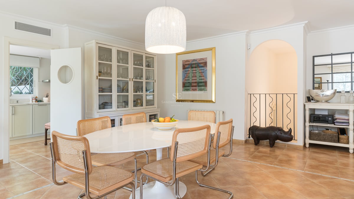 Bright dining room featuring an oval white table, rattan chairs, terracotta floors, and a large display cabinet.