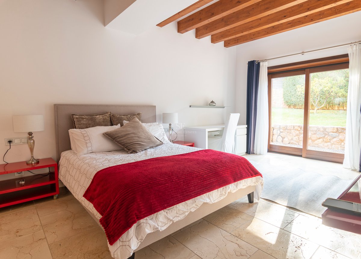 Ljust sovrum med röda detaljer, stengolv, takbjälkar i trä och skjutdörrar i glas mot trädgården.