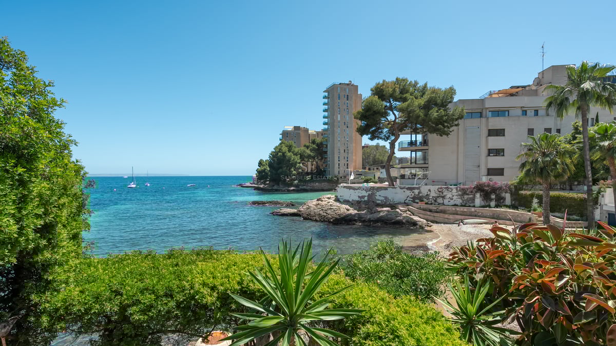 Turkos vik med klippig kust och moderna byggnader inramade av medelhavsgrönska under en klarblå himmel.