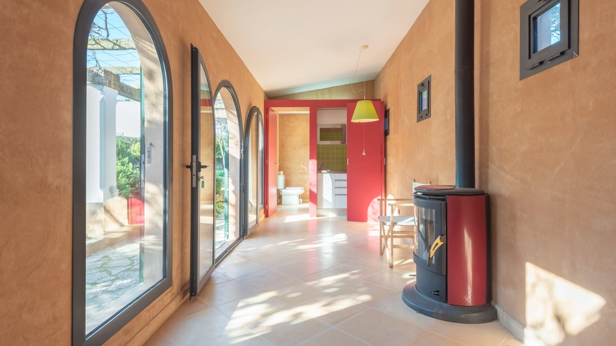 Solbelyst rum med terrakottaväggar, valvade glasdörrar, en röd braskamin och utsikt mot trädgården.