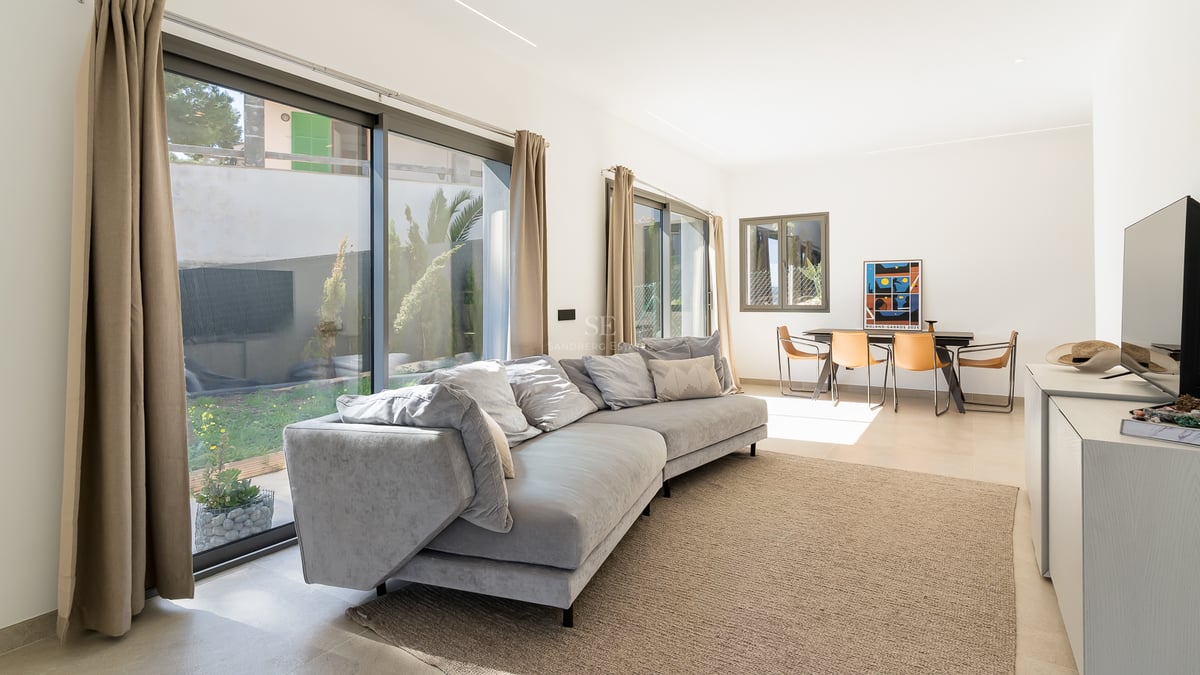 Ljust vardagsrum med grå soffa, stora glaspartier mot trädgården och matplats med läderstolar.