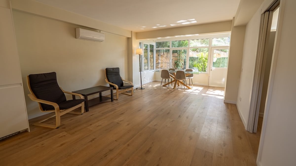 Salón con suelos de madera, grandes ventanales con vistas al jardín, mesa de comedor de cristal y dos sillones negros.