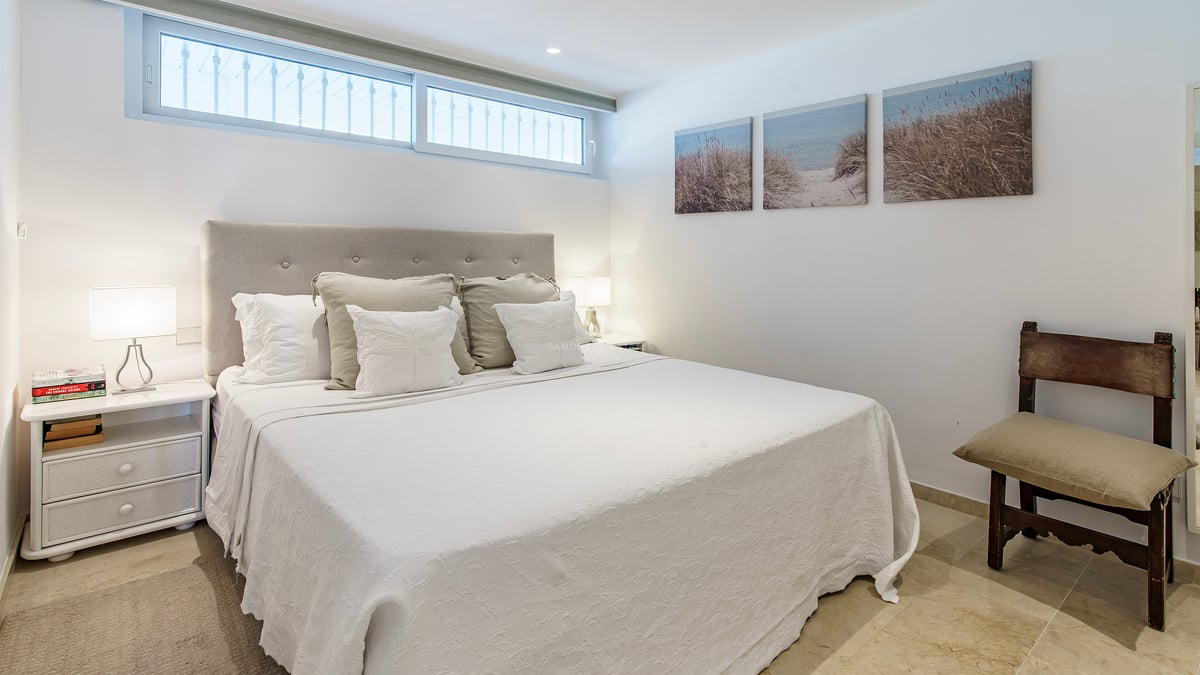 A bright bedroom featuring a large bed with a tufted headboard, white bedding, and coastal artwork on the wall.