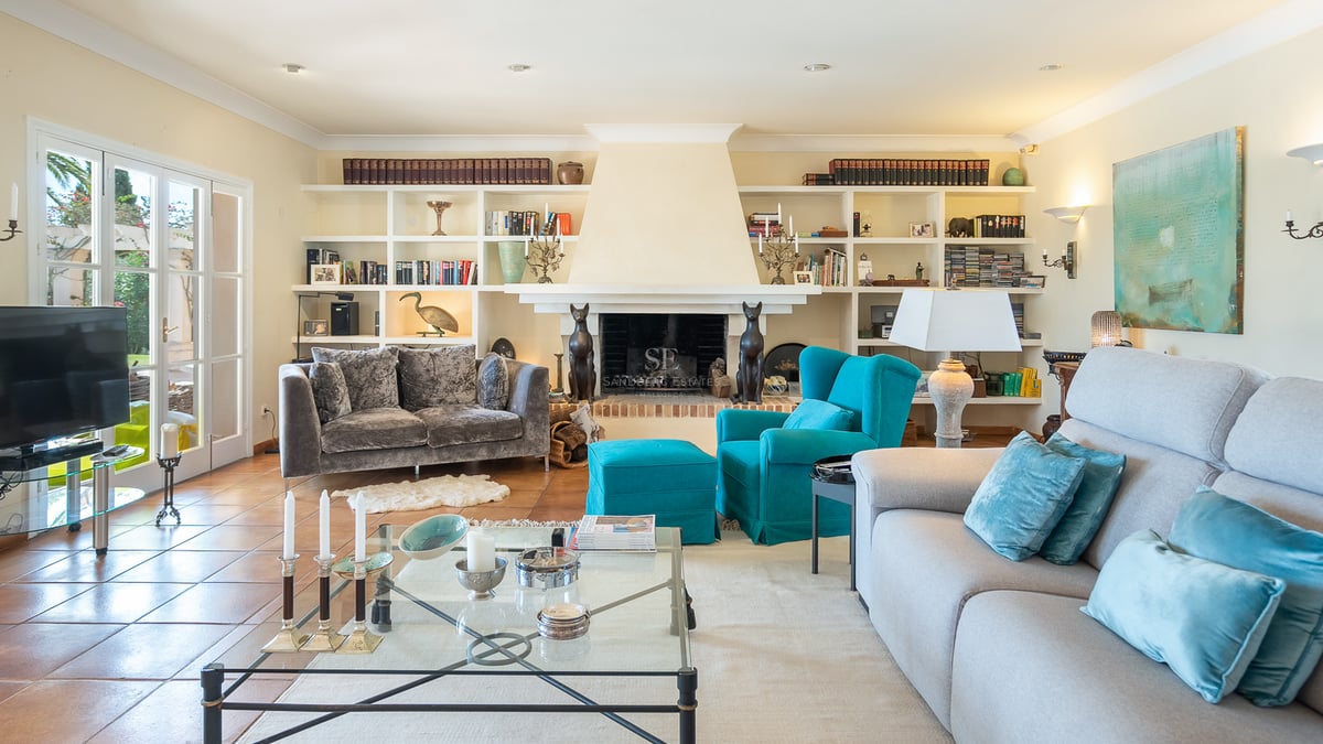 Salon lumineux avec sol en terre cuite, canapés gris, fauteuil turquoise, cheminée et étagères intégrées.