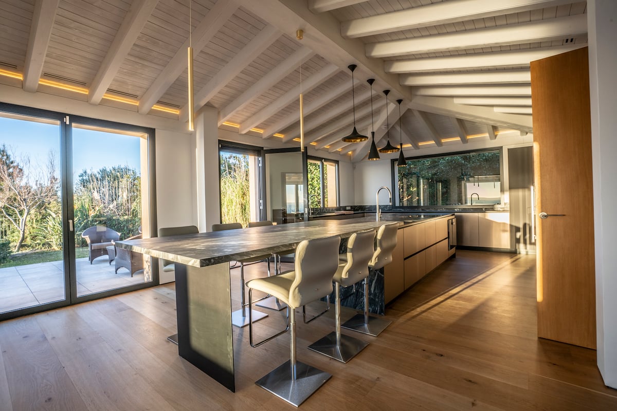 Moderne Küche mit Marmorinsel, weißen Holzbalken und bodentiefen Glasfronten zum Garten.