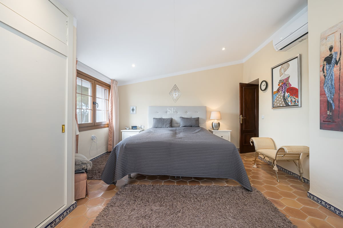 Chambre avec sol en terre cuite, grand lit avec couvre-lit gris, placards intégrés blancs et fenêtre en bois.