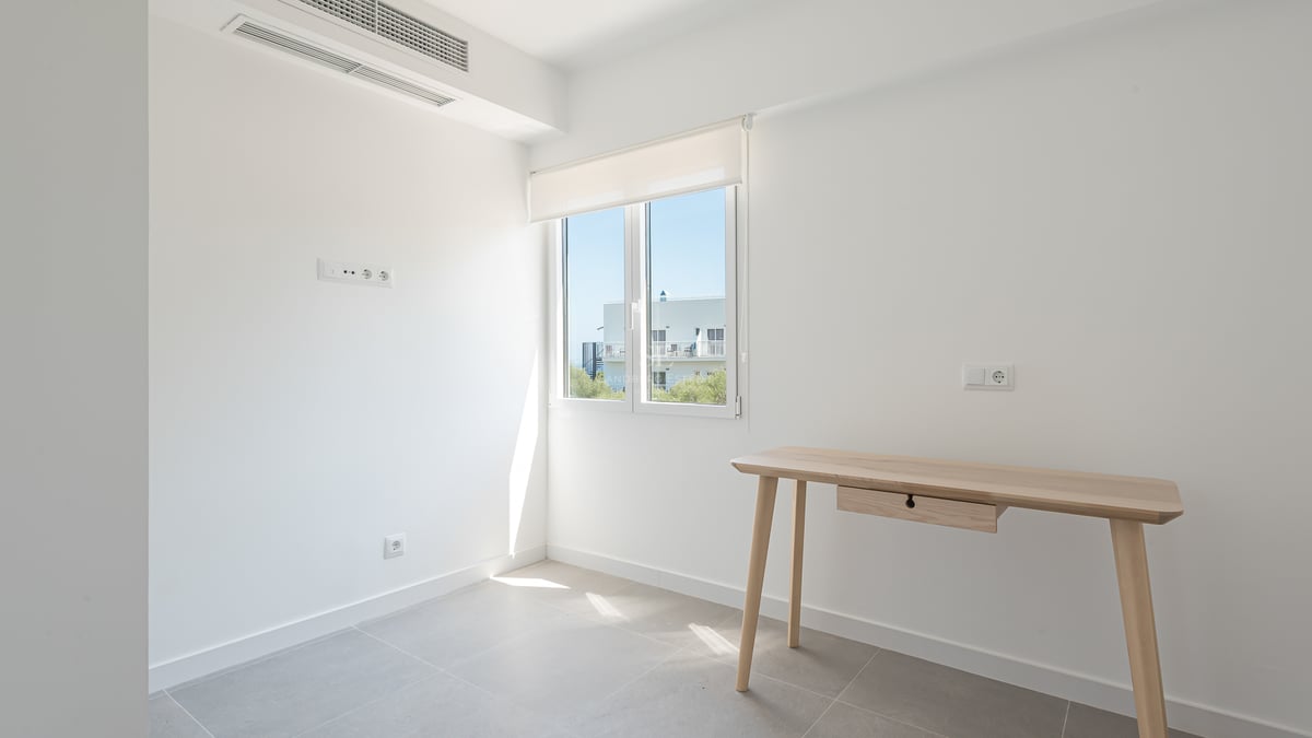 Pièce blanche avec bureau en bois clair, sol en carrelage gris et fenêtre offrant une lumière vive.