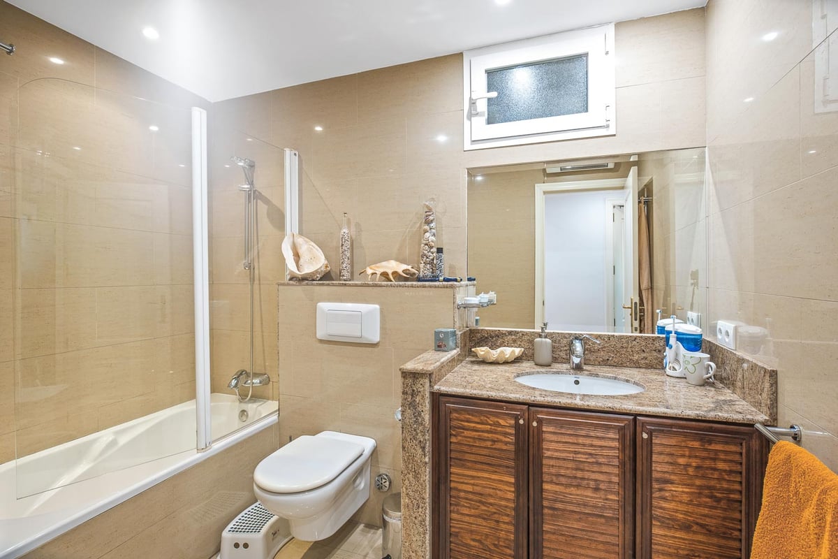 Badezimmer mit Badewanne, Glaswand, dunklem Holz-Waschtisch, Granitplatte und sandfarbenen Fliesen.