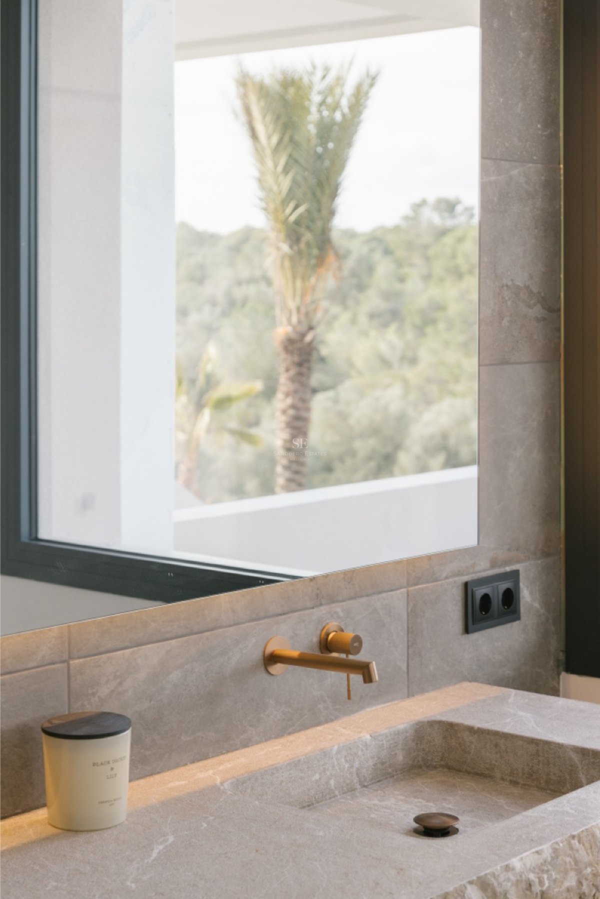 Nahaufnahme eines integrierten Natursteinwaschbeckens mit Messingarmatur und Spiegel mit Palmenreflexion.