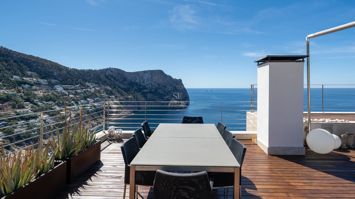 Modern rooftop terrace with a large dining table overlooking the sea and rugged coastline under a clear blue sky.