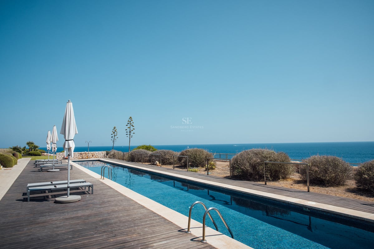Longue piscine rectangulaire avec terrasse en bois sombre, parasols blancs et vue imprenable sur la mer azur.