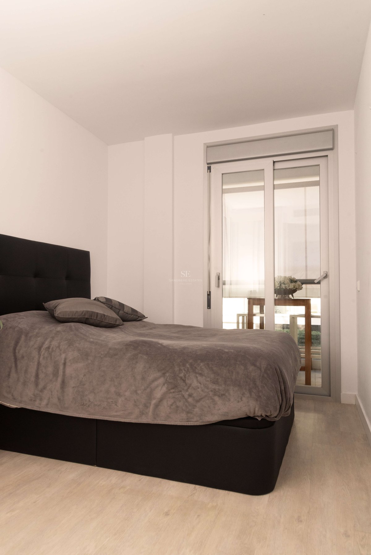 Schlafzimmer mit grauem Bett, schwarzem Kopfteil, hellem Holzboden und Glasschiebetür zur Terrasse.