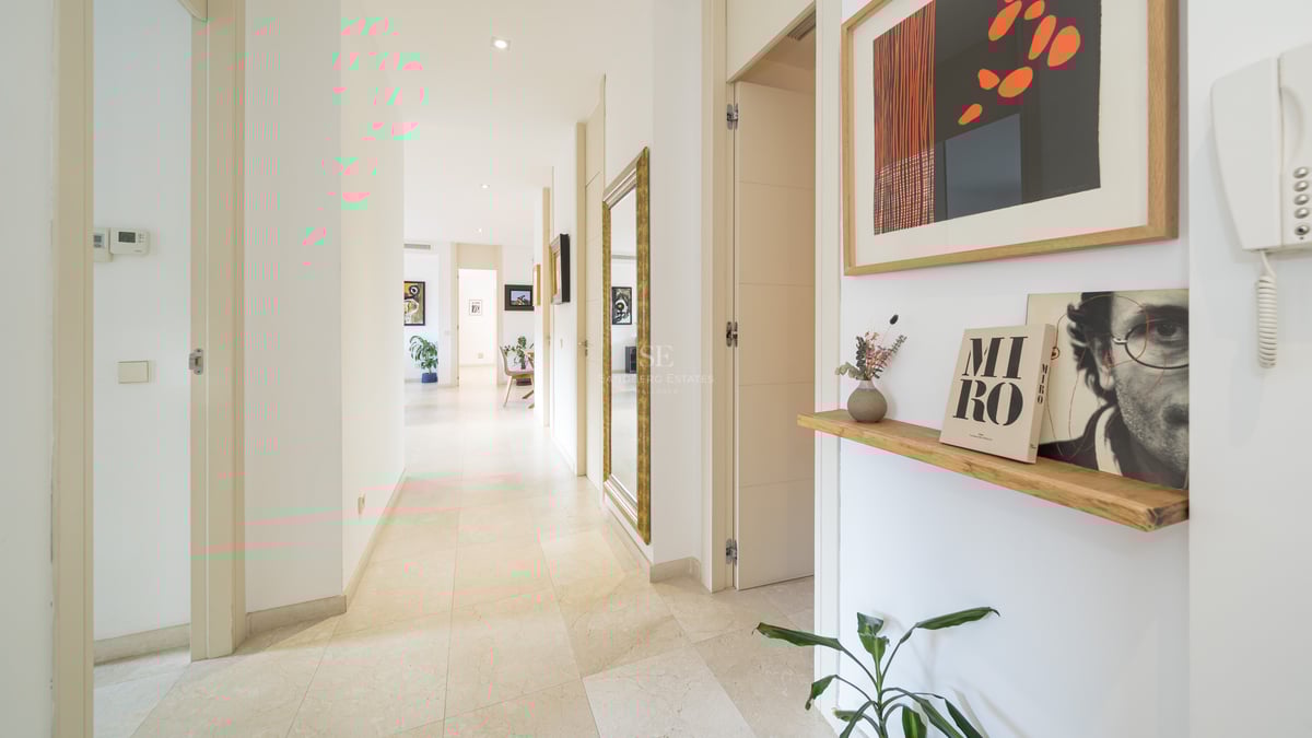 Lång vit korridor med stengolv, en stor guldinramad spegel och färgglada konstverk.