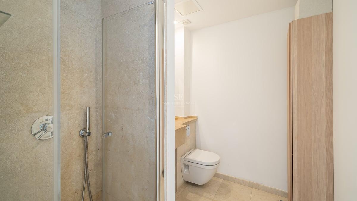 Badrum med glasdusch, vägghängd toalett och beiga stenväggar.