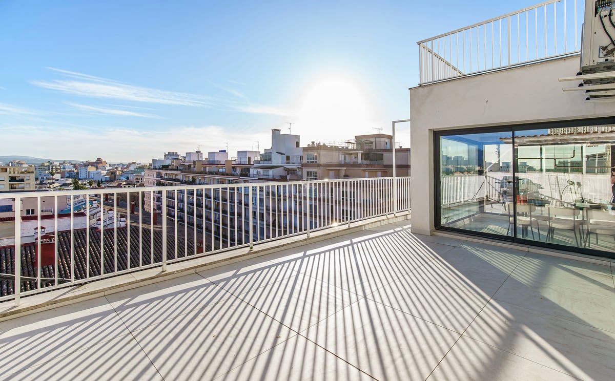 Vaste terrasse de penthouse avec carrelage et garde-corps blancs, offrant une vue sur la ville sous un soleil éclatant.