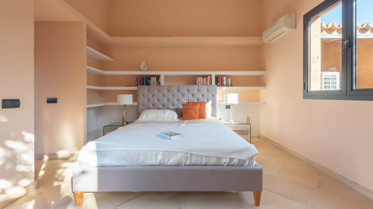 Ljust sovrum med grå tuftad sänggavel, vita sängkläder, inbyggda hyllor och terrakottagolv.