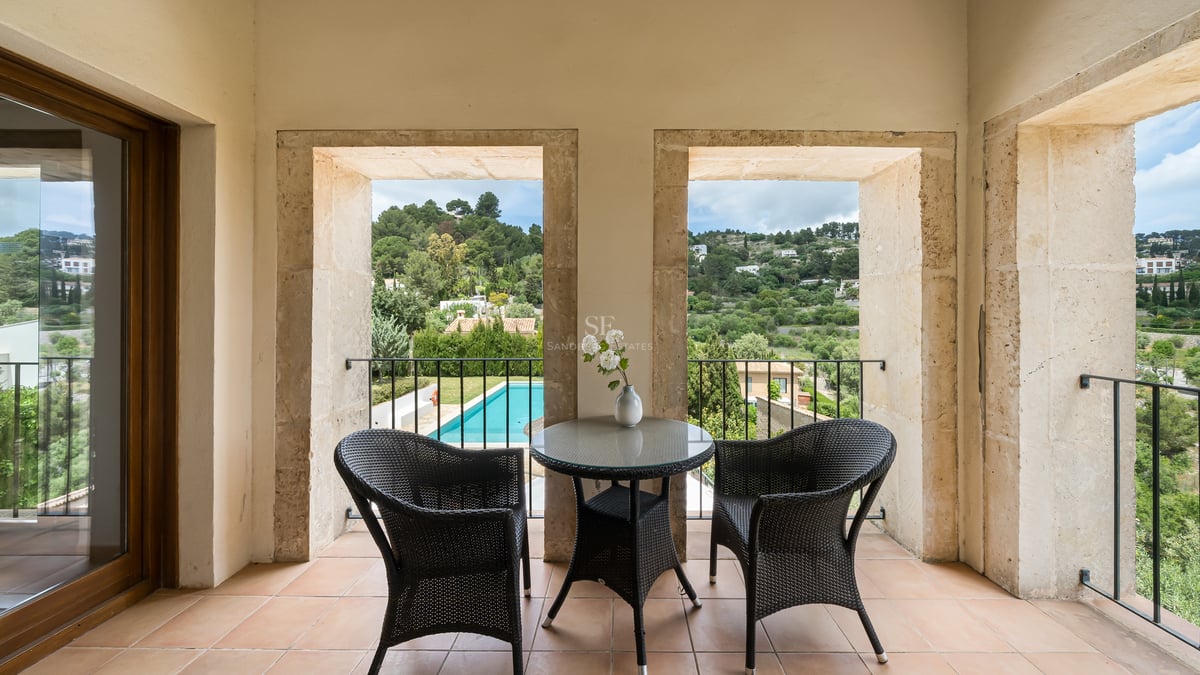 Deux chaises en osier et une table sur une terrasse encadrée de pierre avec vue sur une piscine et des collines vertes.