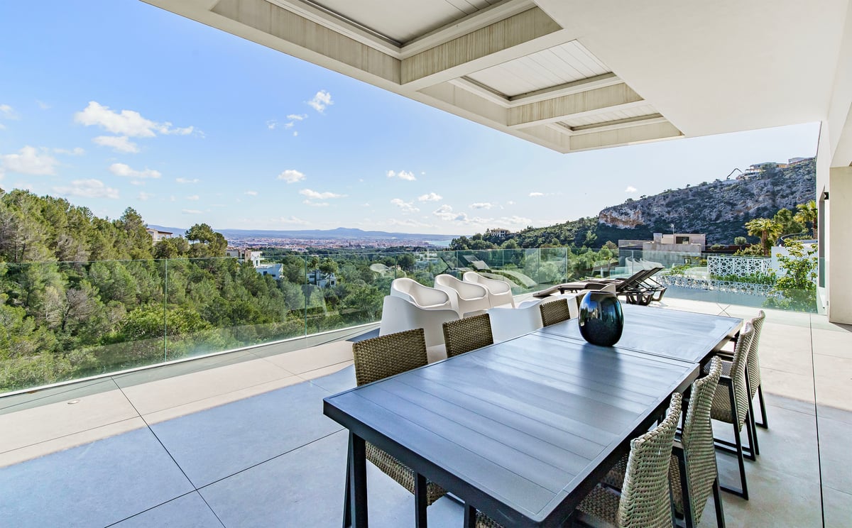 Terrasse moderne avec table à manger et garde-corps en verre surplombant une forêt et la ville au loin.