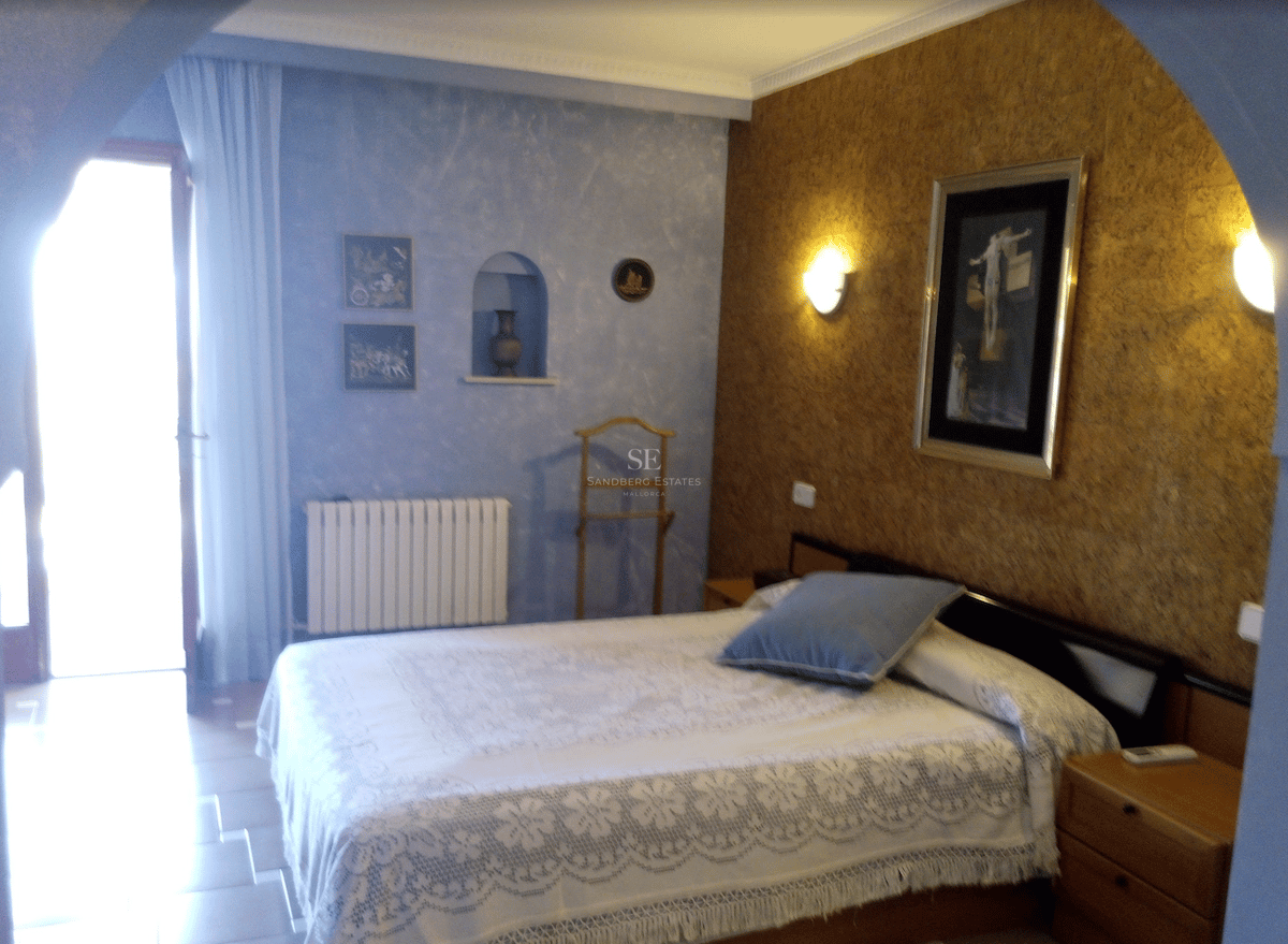 Dormitorio con colcha de encaje blanco, pared azul con textura y luz natural desde una puerta lateral.