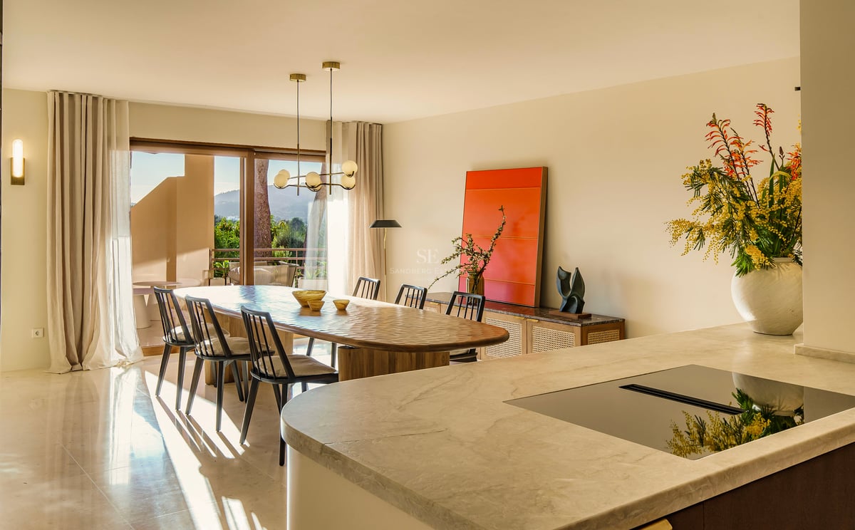 Ovaler Holzesstisch mit schwarzen Stühlen vor großen Glasfronten und einer Marmor-Kücheninsel im Vordergrund.