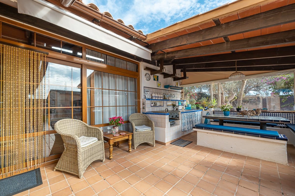 Terrasse couverte comprenant une cuisine d'été, des poutres en bois, un sol en terre cuite et des fauteuils en osier.